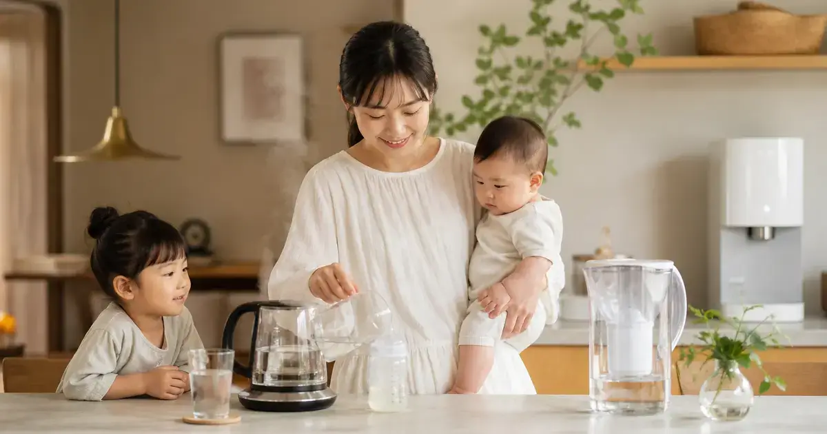 赤ちゃんを抱いた母親がキッチンでミルクを準備し、そばで子どもが見守る家庭の水まわりのイメージ。煮沸用のケトルや浄水ポットが置かれている。
